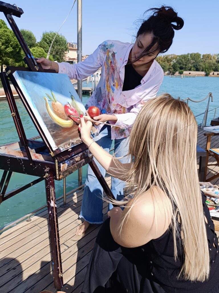 Workshop di pittura creativa in barca durante un’attività di Team Building a Venezia, con partecipanti che dipingono un frutto su tela vicino alla laguna.