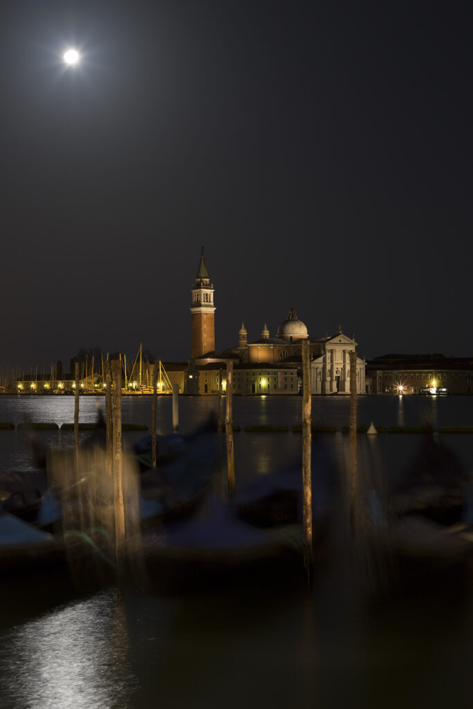 Veduta notturna di Venezia con la luna piena su San Giorgio Maggiore e gondole in ombra, atmosfera misteriosa e suggestiva.